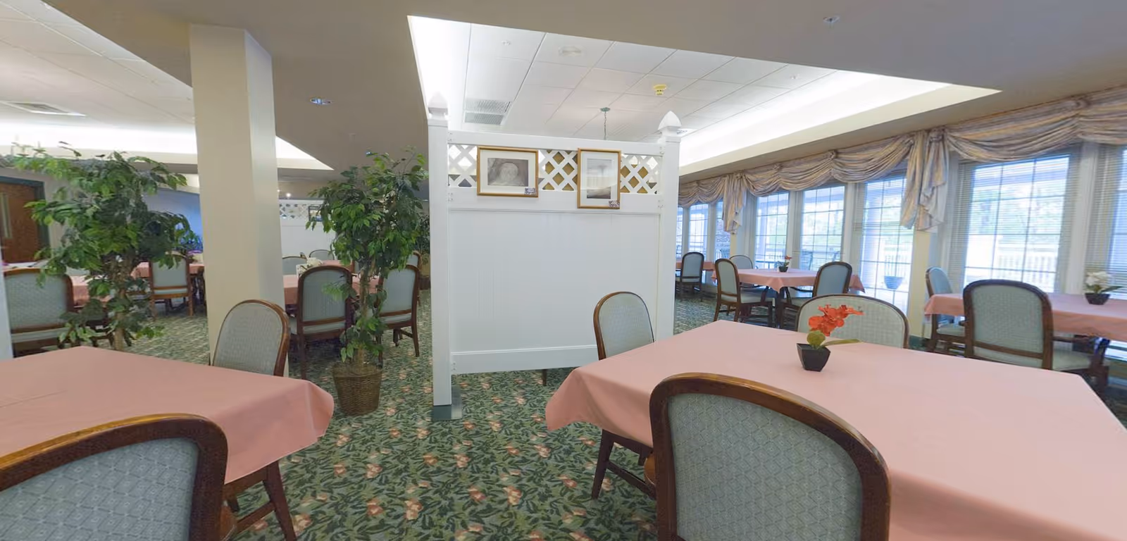 A dining room with multiple tables covered in pink tablecloths and surrounded by cushioned chairs. The room features large windows with draped curtains allowing natural light to enter. There are potted plants and a white partition with framed pictures hanging on it in the center of the room. The carpet has a floral pattern.