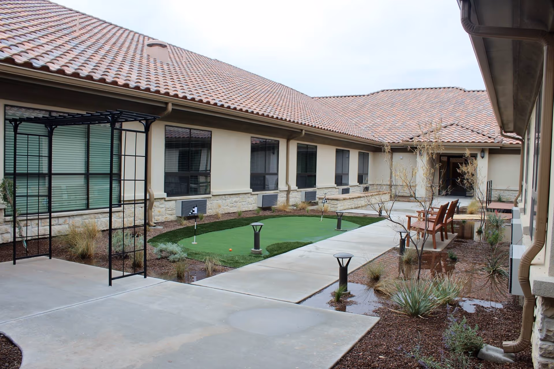 Outdoor courtyard area at VibraLife El Paso now Ignite Medical Resorts featuring a small putting green with golf flags, concrete walkways, desert landscaping with plants and shrubs, wooden benches, and a building with tiled roof and multiple windows surrounding the courtyard.