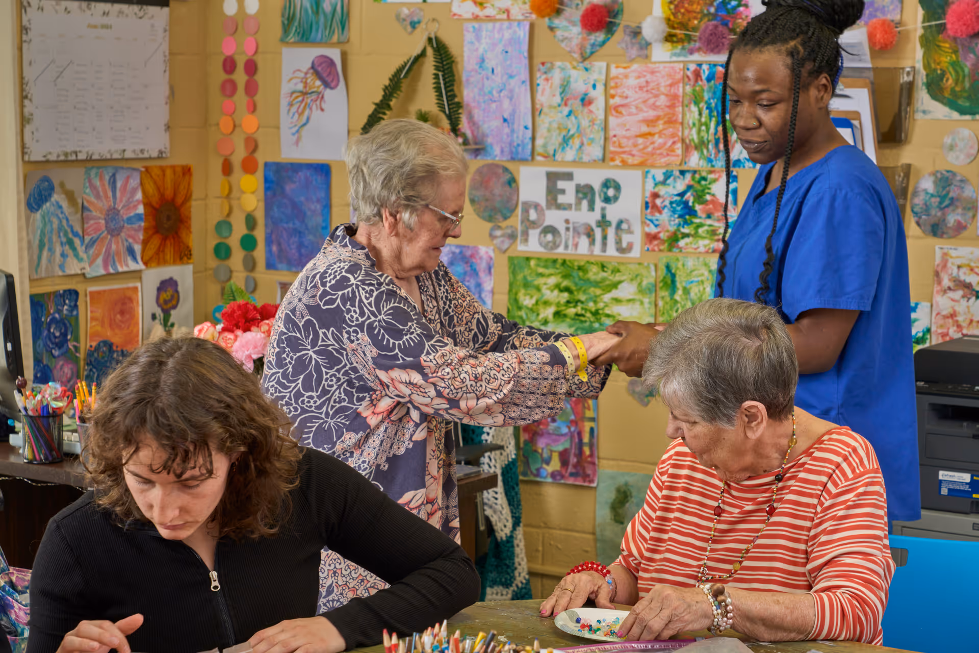 A group of people engaged in arts and crafts activities in a colorful room decorated with various paintings and artwork. Two elderly women and a younger woman are seated at a table working on crafts, while a caregiver in blue scrubs holds hands with one of the elderly women. The wall behind them displays the name 'Eno Pointe' among the artwork.