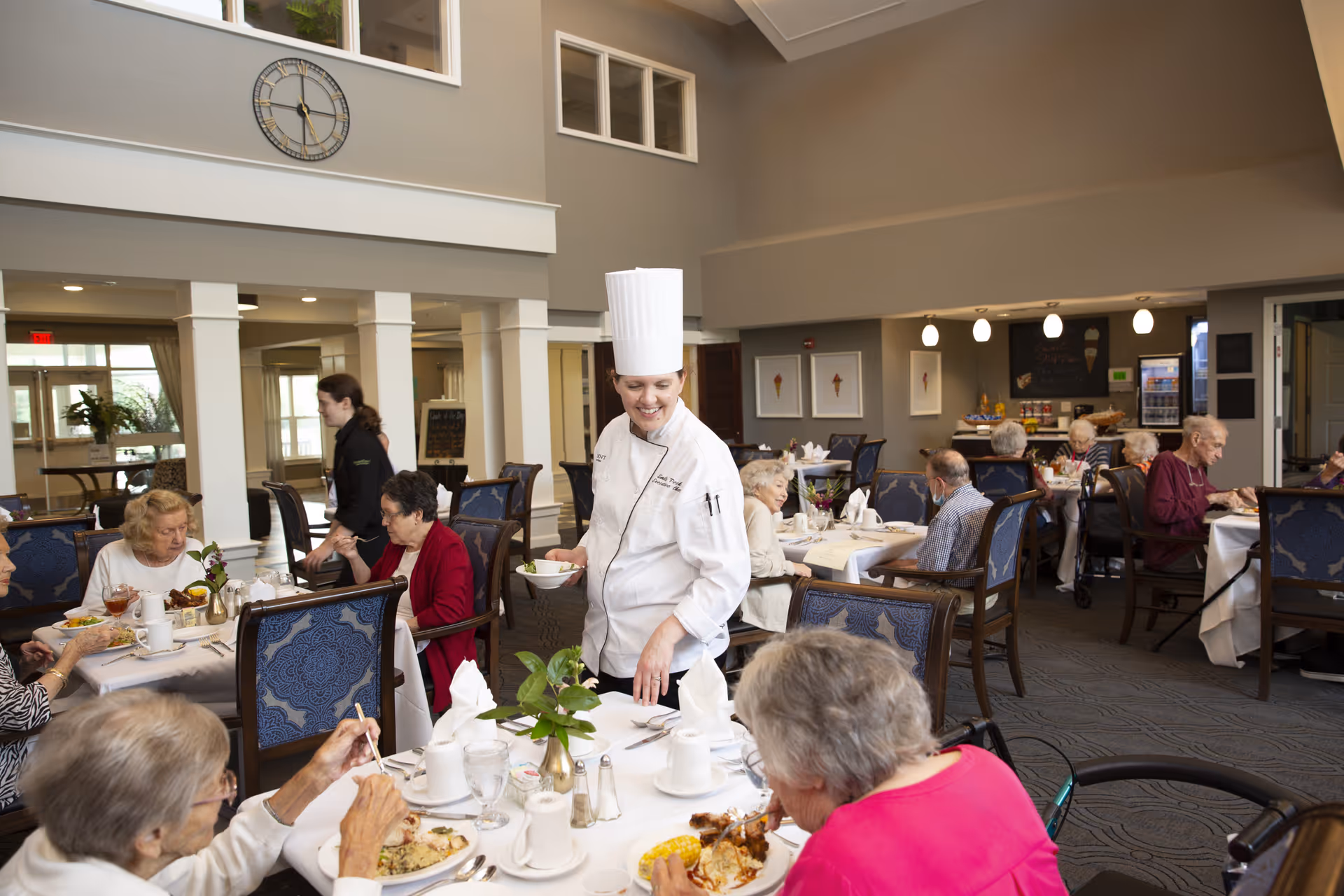 A dining room in a senior living facility with elderly residents seated at tables enjoying their meals. A chef in a white uniform and tall hat is serving food and smiling. The room has high ceilings, large windows, and elegant decor with blue upholstered chairs and white tablecloths.