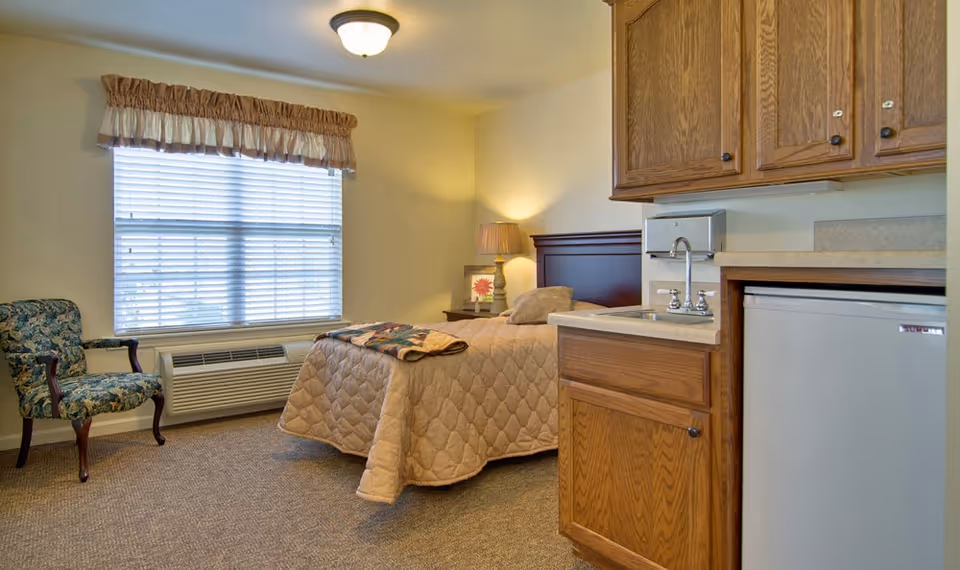 A cozy senior living bedroom with a single bed covered in a beige quilt, a wooden headboard, a bedside table with a lamp and a framed flower picture, a floral upholstered armchair near a window with blinds and a valance, and a small kitchenette area with wooden cabinets, a sink, and a mini refrigerator.