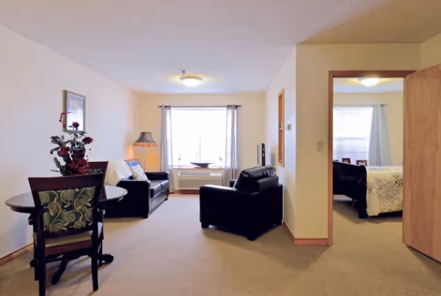 A cozy living area with two black leather chairs and a small round dining table with two chairs. A window with light curtains lets in natural light. To the right, an open doorway reveals a bedroom with a bed covered in a floral bedspread.