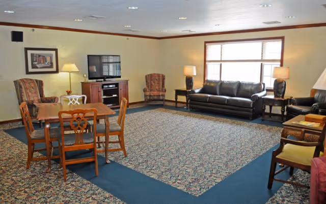 A spacious living room with patterned carpet and beige walls. The room features a wooden table with four chairs, two upholstered armchairs, a black leather sofa, a black leather armchair, two side tables with lamps, a TV on a wooden stand, and a large window with blinds letting in natural light.