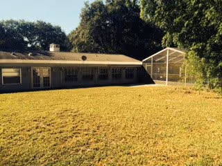 Single-story building with multiple windows and a screened-in porch on the right side, surrounded by a large grassy yard and trees in the background under a clear sky.