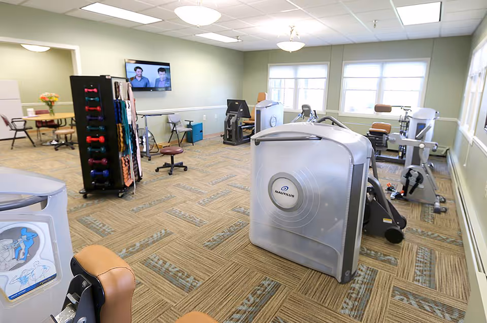 Bright senior fitness room with exercise machines, a dumbbell rack, chairs, and a wall-mounted TV under large windows.