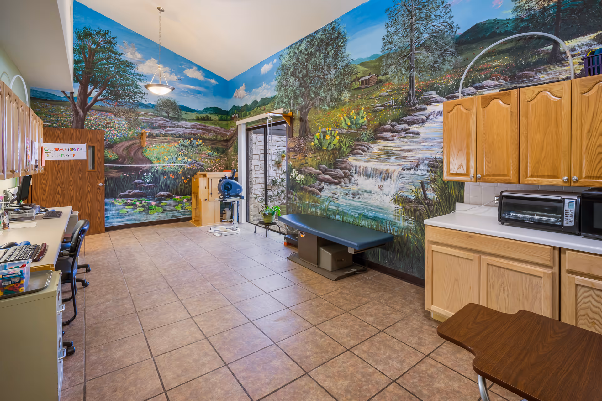 A spacious room with tiled flooring and a high ceiling, featuring a large mural of a natural landscape with trees, a stream, and mountains on the walls. The room contains wooden cabinets, a countertop with a microwave and toaster oven, a medical examination table, office chairs, and desks with computer equipment. There is also a door and a window letting in natural light.