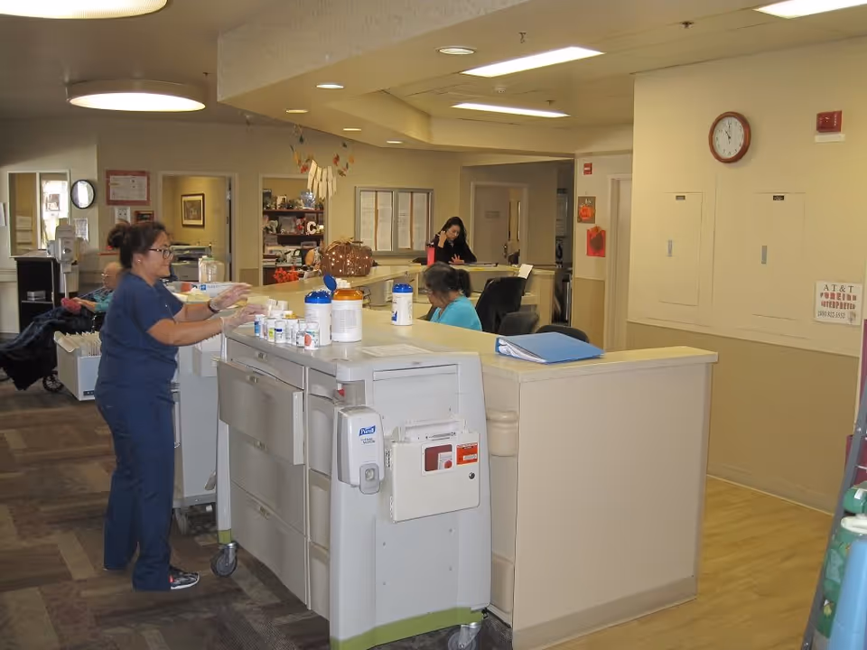 A nurse in blue scrubs is preparing medication at a nursing station in a senior living facility. Behind the station, two women are seated and working, one on the phone and the other looking down. The area is well-lit with overhead lights, and there are various medical supplies and paperwork on the counter. The environment appears clean and organized with a clock on the wall and some decorations.