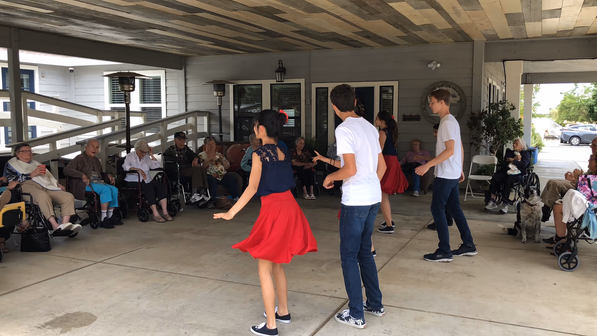 A group of young people dancing under a covered outdoor area while elderly residents sit around watching. The elderly are seated in chairs and wheelchairs along the perimeter, and there is a dog sitting near one of the residents. The setting appears to be a senior living facility with a concrete floor and a wooden ceiling.