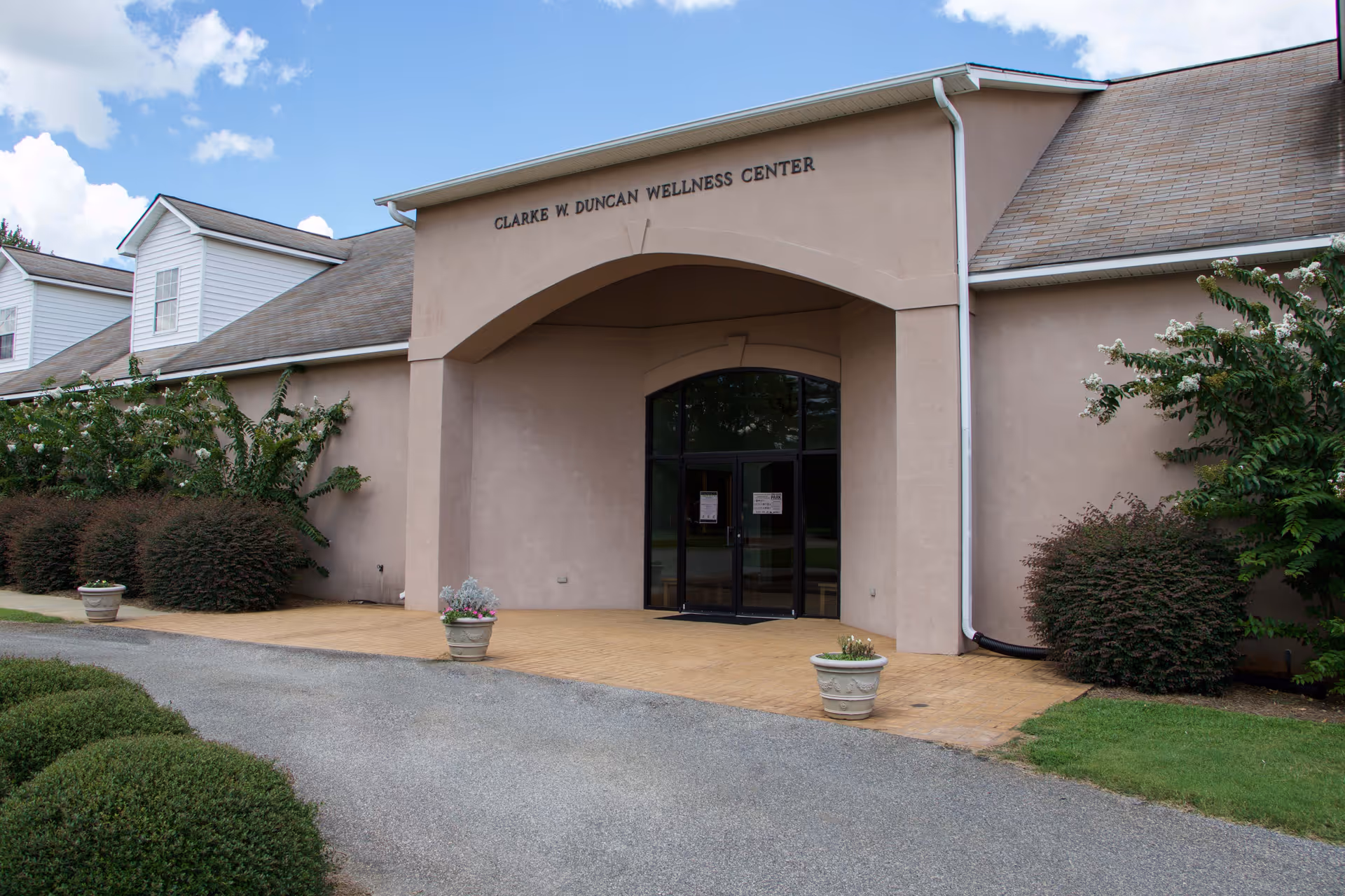Exterior view of the Clarke W. Duncan Wellness Center building with a beige facade, arched entrance, glass double doors, and surrounding greenery including bushes and potted plants.
