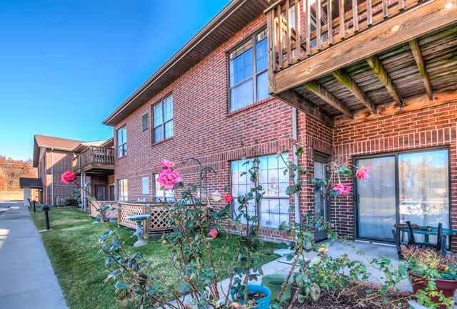 Exterior view of a brick senior living facility building with balconies and large windows. There are pink roses and other plants in the garden area in front of the building, with a sidewalk running alongside the building under a clear blue sky.