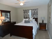 A bedroom with a dark wooden bed frame and white bedding, two patterned pillows, a matching dark wooden dresser with a mirror, a nightstand with a lamp, beige walls, carpeted floor, and a ceiling fan with a light. Two windows with patterned curtains are behind the bed.