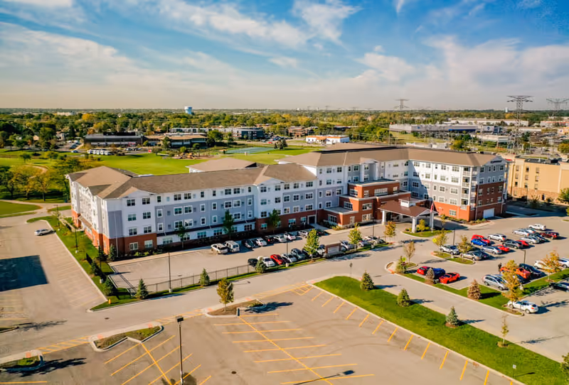 Aerial view of a large senior living facility building with a parking lot in front, surrounded by greenery and other buildings under a partly cloudy sky.