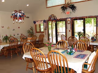 A dining room with several wooden tables and chairs arranged neatly. The tables are covered with white tablecloths and decorated with small floral centerpieces. The room has large windows and glass doors letting in natural light, with patriotic decorations hanging from the ceiling and walls. There are plants placed around the room and an exit sign above the door.