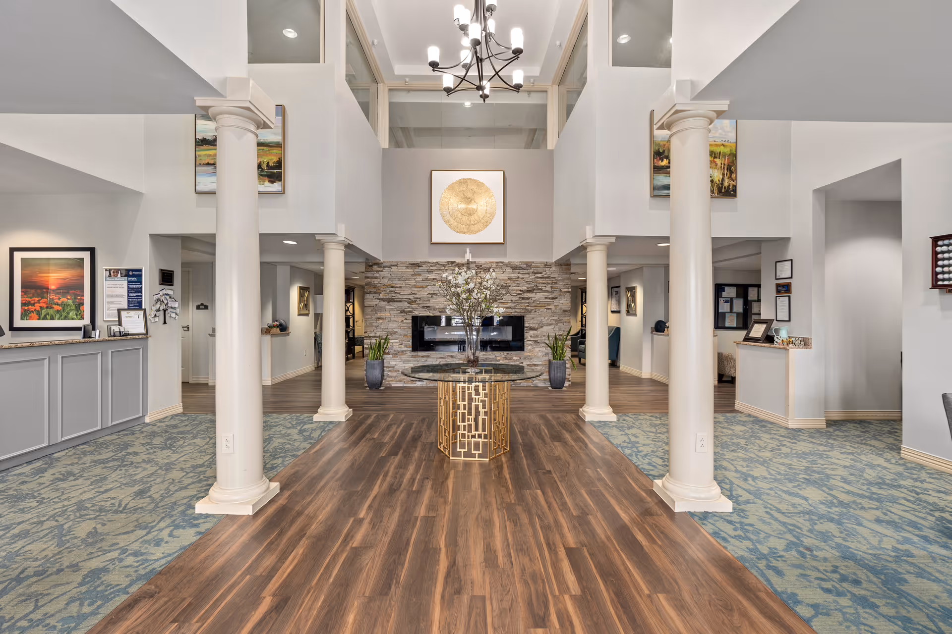 Spacious and elegant interior lobby area with high ceilings, four white columns, a chandelier, a round glass table with a decorative vase in the center, wood flooring in the middle, and patterned carpet on the sides. The back wall features a stone fireplace with a large circular gold artwork above it. There are framed pictures on the walls and reception desks on both sides.