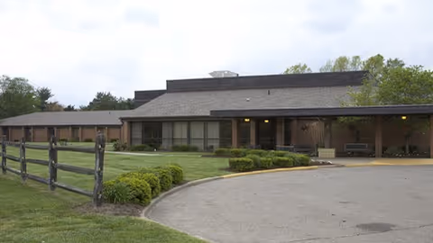 Single-story brick senior living building with a covered drive-up entrance, curved driveway, lawn and split-rail fence.