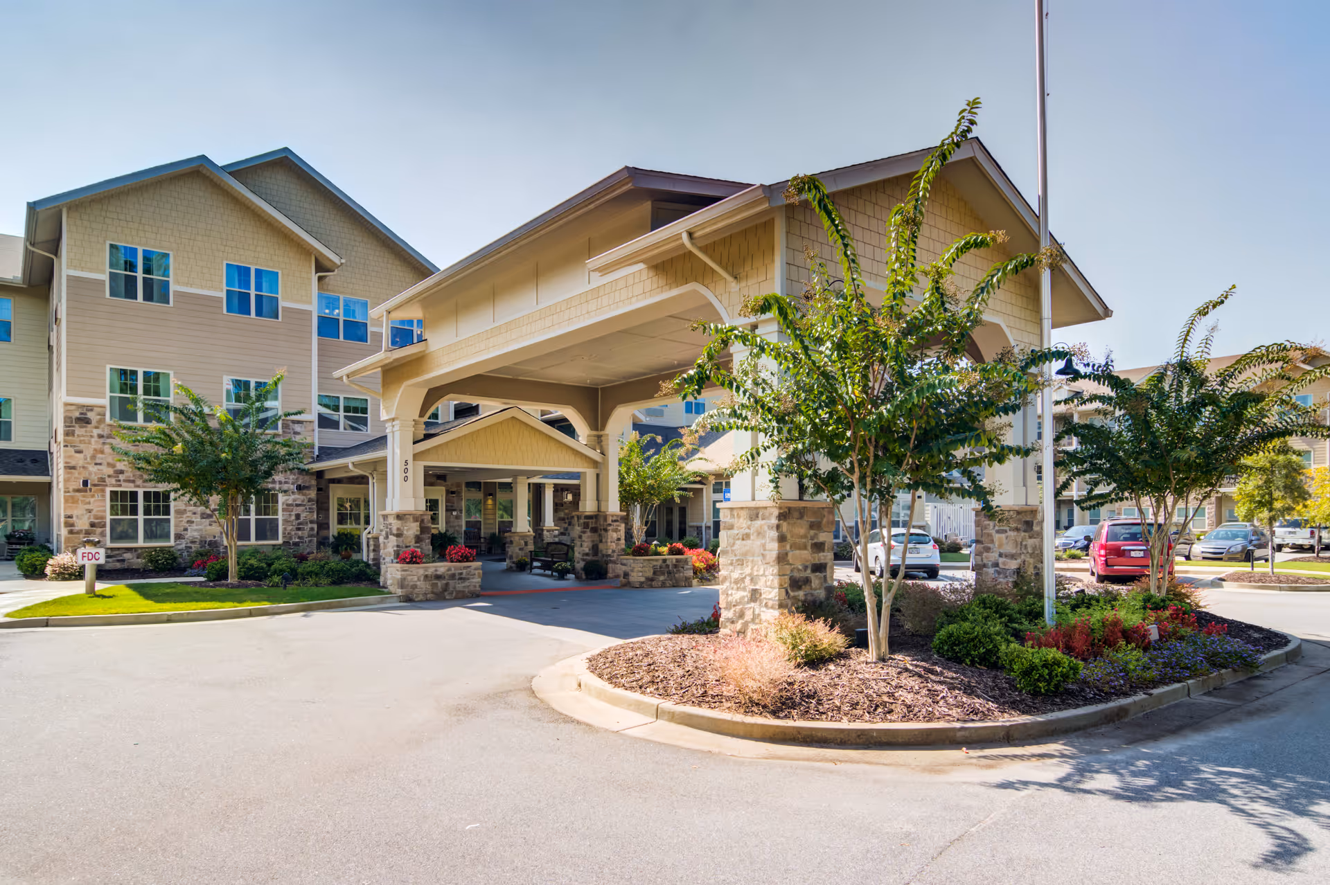 Exterior view of Sanford Estates Gracious Retirement Living facility showing a covered entrance with stone pillars, landscaped greenery, trees, and parked cars under a clear blue sky.