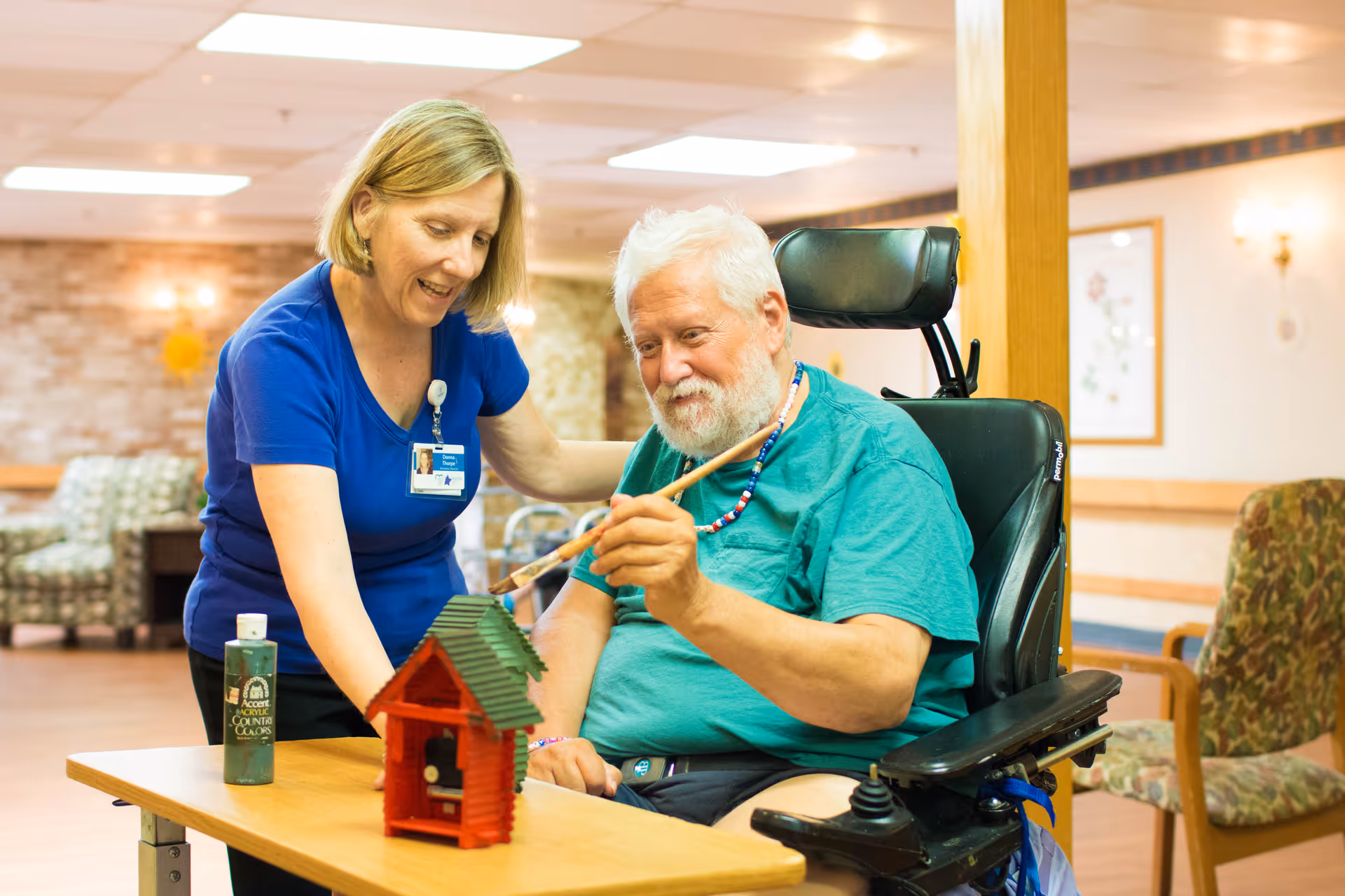 A senior man in a wheelchair is painting a small wooden birdhouse with the assistance of a female caregiver in a blue shirt. They are in a well-lit room with comfortable chairs and warm decor.