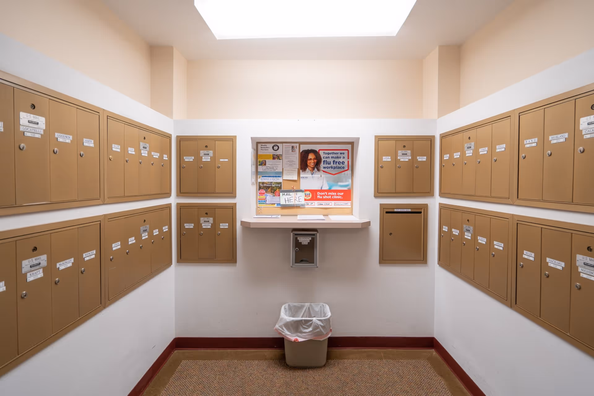 A mailroom with multiple beige mailboxes mounted on three walls. In the center, there is a bulletin board with various notices, including a poster about making a flu-free workplace and a sign indicating where to place mail. Below the bulletin board is a small counter and a locked drop box labeled 'Resident'. A trash can with a plastic liner is placed on the floor beneath the counter.