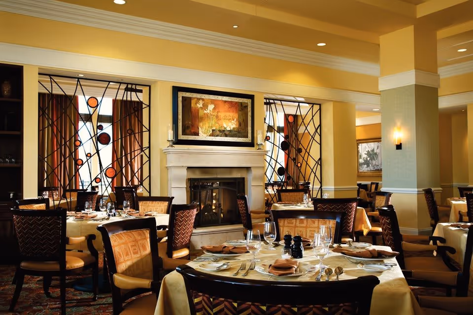 A warmly lit dining room with multiple tables set with plates, glasses, silverware, and napkins. The room features a fireplace with a framed artwork above it, decorative metal room dividers, and upholstered chairs. The walls are painted in soft yellow tones, and there are recessed ceiling lights providing illumination.