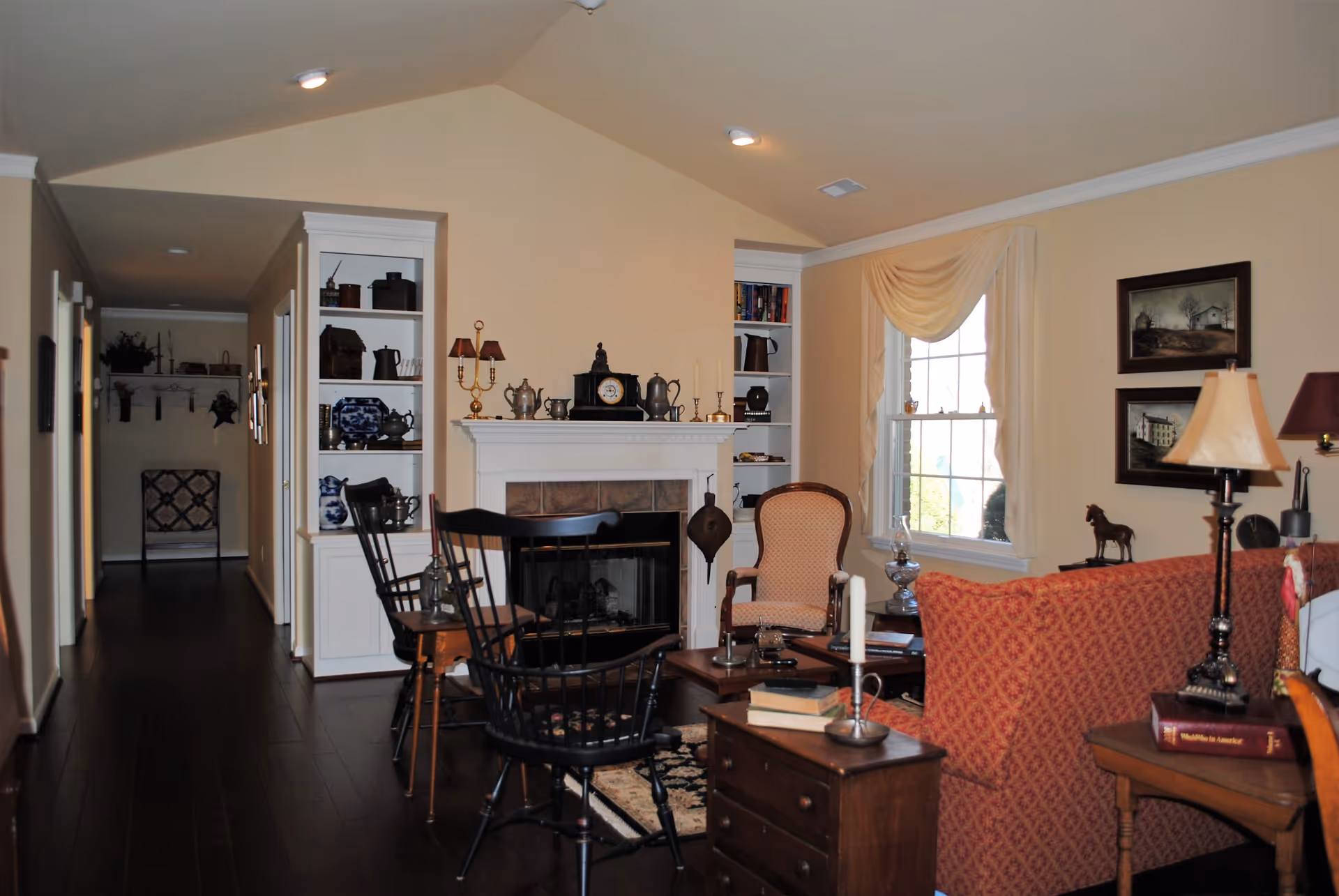 A cozy living room with a fireplace in the center, flanked by built-in shelves filled with decorative items. There are two black wooden chairs and an upholstered armchair around a wooden coffee table. A red patterned sofa is partially visible, along with a side table holding a lamp and books. The room has a large window with cream-colored drapes, framed artwork on the walls, and dark wooden flooring.
