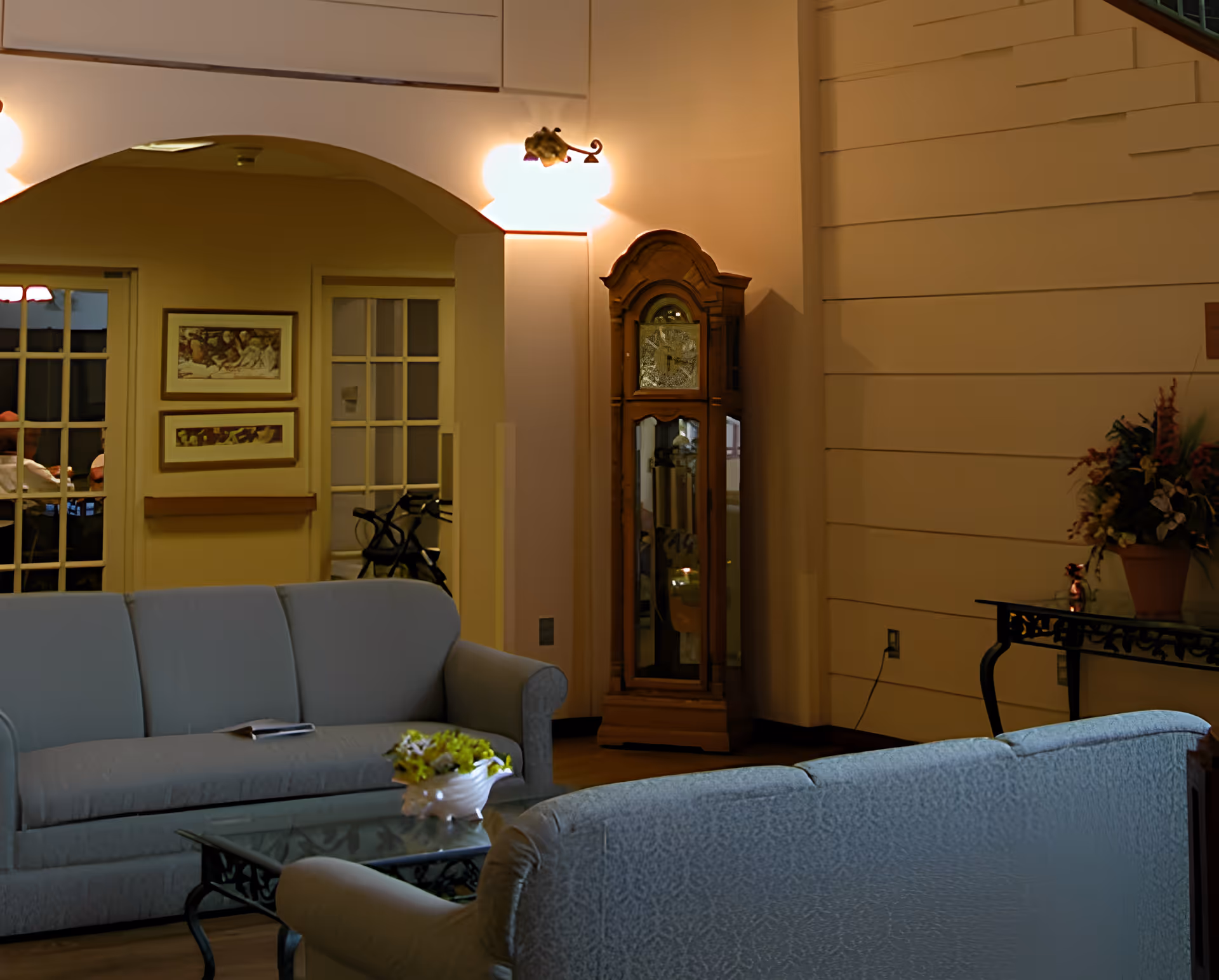 A cozy living room area with two light blue sofas facing each other, a glass coffee table with a decorative flower arrangement, a wooden grandfather clock against the wall, and a side table with a flower pot. The room has warm lighting and an archway leading to another room with glass-paneled doors.