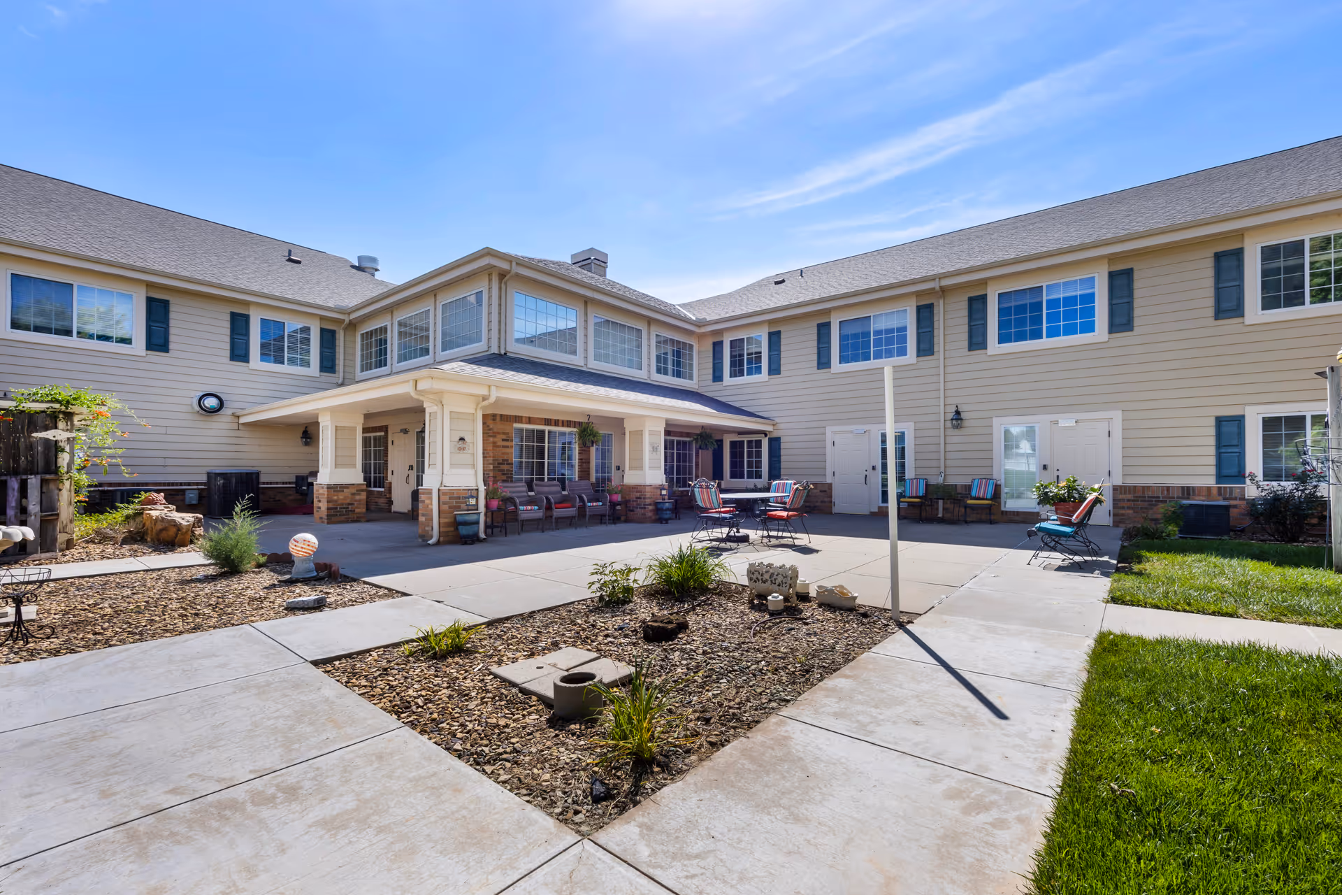 Outdoor courtyard area of a senior living facility with beige siding and brick accents. The courtyard features concrete walkways, a small garden bed with plants and decorative items, several chairs with colorful cushions, and a table. The building has multiple windows and a covered porch area with additional seating.