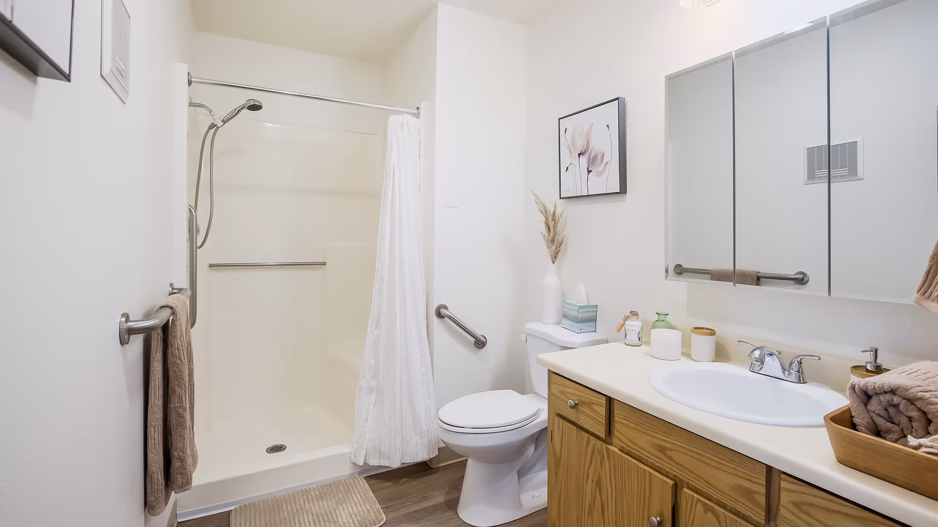 A clean and bright bathroom featuring a walk-in shower with a white curtain, a toilet with a decorative vase and tissue box on top, and a wooden vanity with a white sink and various toiletries. There is a large mirror above the sink and a towel hanging on a rail next to the shower.