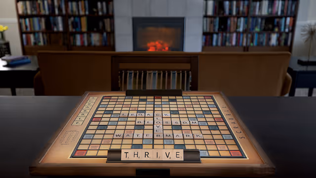 A Scrabble board on a table with the words LEGACY, CLOVER, BLOSSOM, WATERMARK, and THRIVE spelled out. In the background, there is a cozy living room with a fireplace, bookshelves filled with books, and a brown sofa.