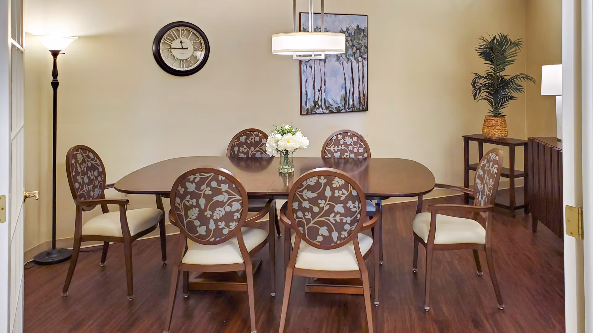 A dining room with a wooden table surrounded by six chairs with floral patterned backs and cream-colored seats. A vase with white flowers is placed in the center of the table. The room has wooden flooring, a floor lamp on the left, a wall clock above the table, a painting on the wall, and a small table with a potted plant on the right side.