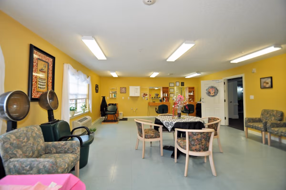 A bright room with yellow walls featuring salon hair dryers, floral upholstered chairs, a round table with a lace tablecloth and a flower centerpiece, and a vanity area with mirrors and chairs. The room has fluorescent ceiling lights and a window with white curtains.