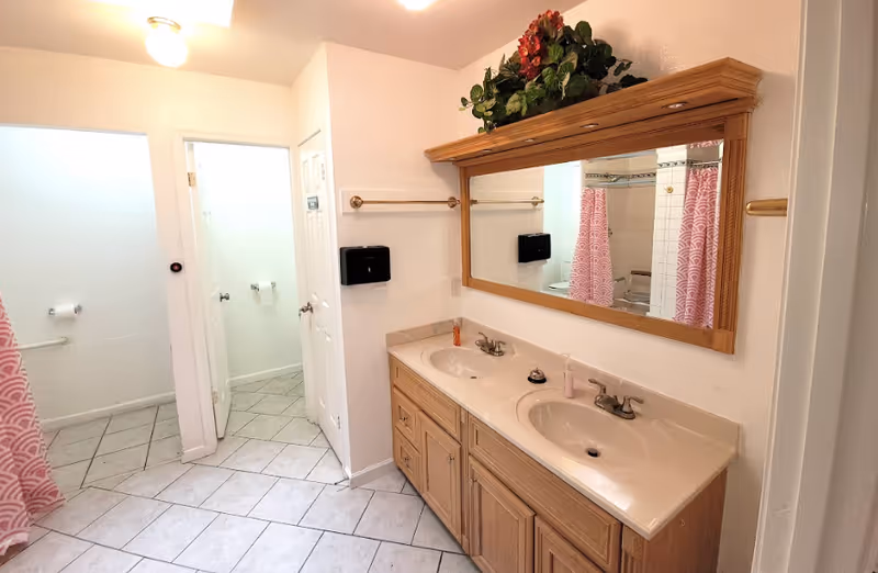 Bright bathroom with a double-sink vanity, large mirror, tiled floor, and a shower with a pink curtain.