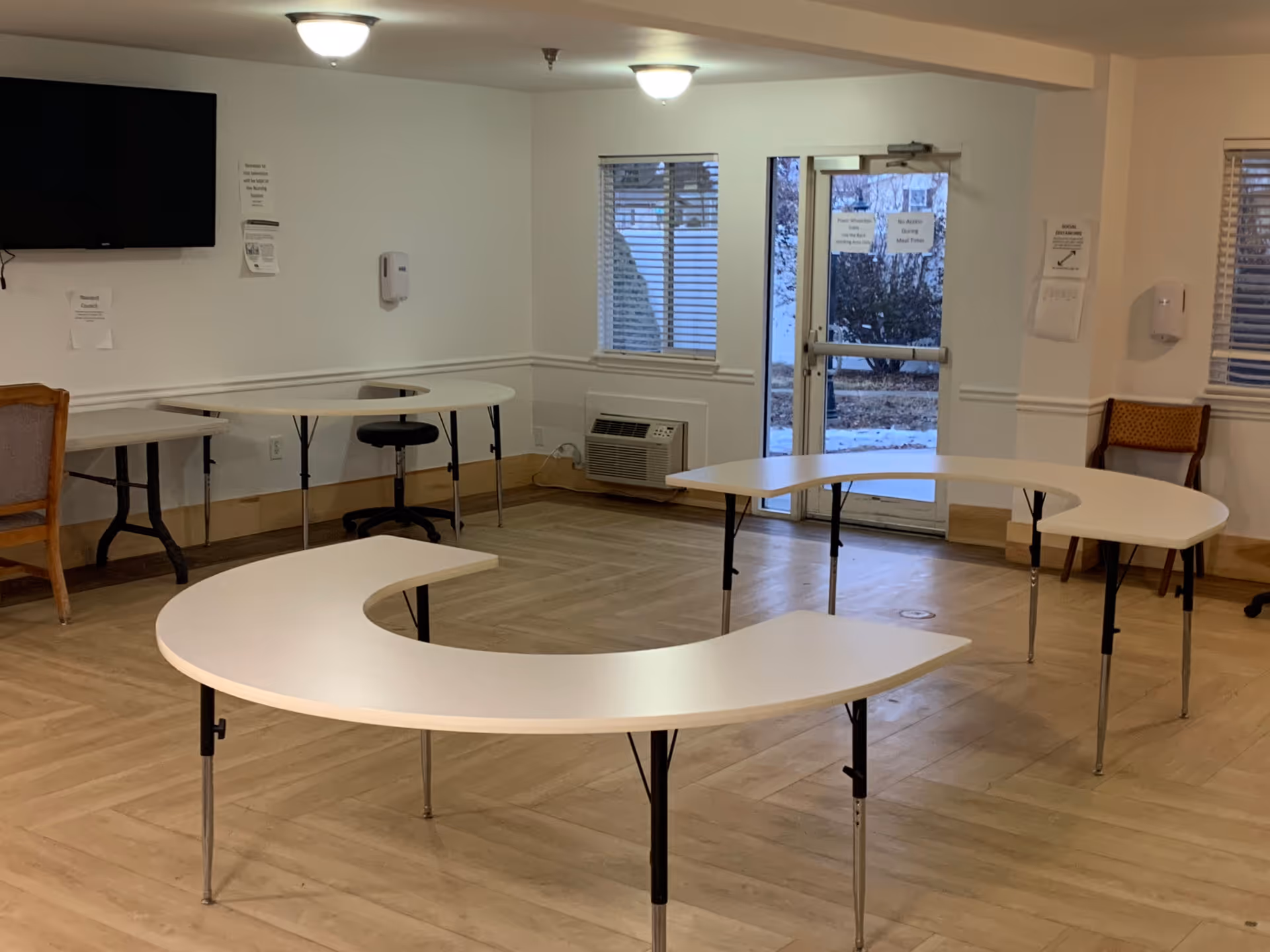 Interior room with light wood flooring and white walls, featuring two white semi-circular tables arranged in a U-shape, a black swivel chair, a wooden chair, a wall-mounted TV, windows with blinds, and a glass door leading outside.
