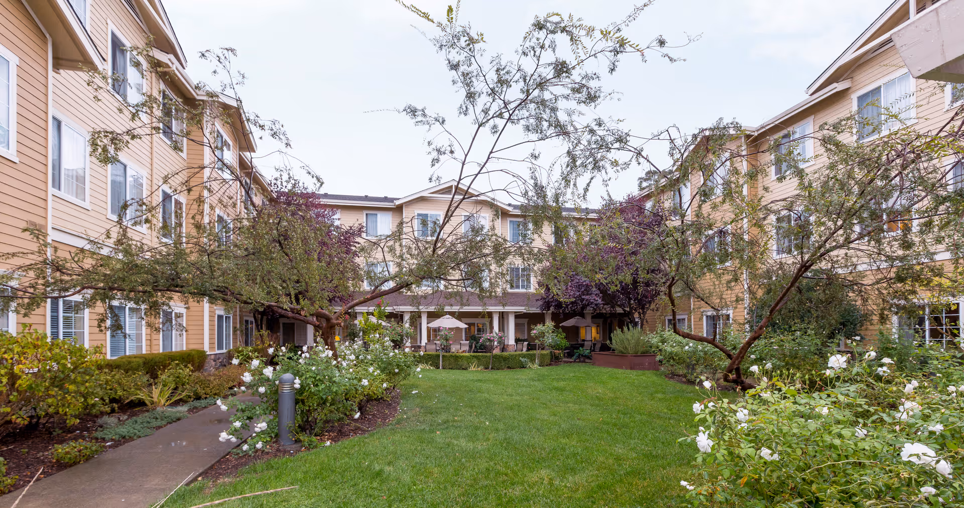 Outdoor courtyard area of Arbol Residences of Santa Rosa featuring a green lawn, flowering bushes, trees, and a paved walkway. The courtyard is surrounded by a three-story beige building with multiple windows.