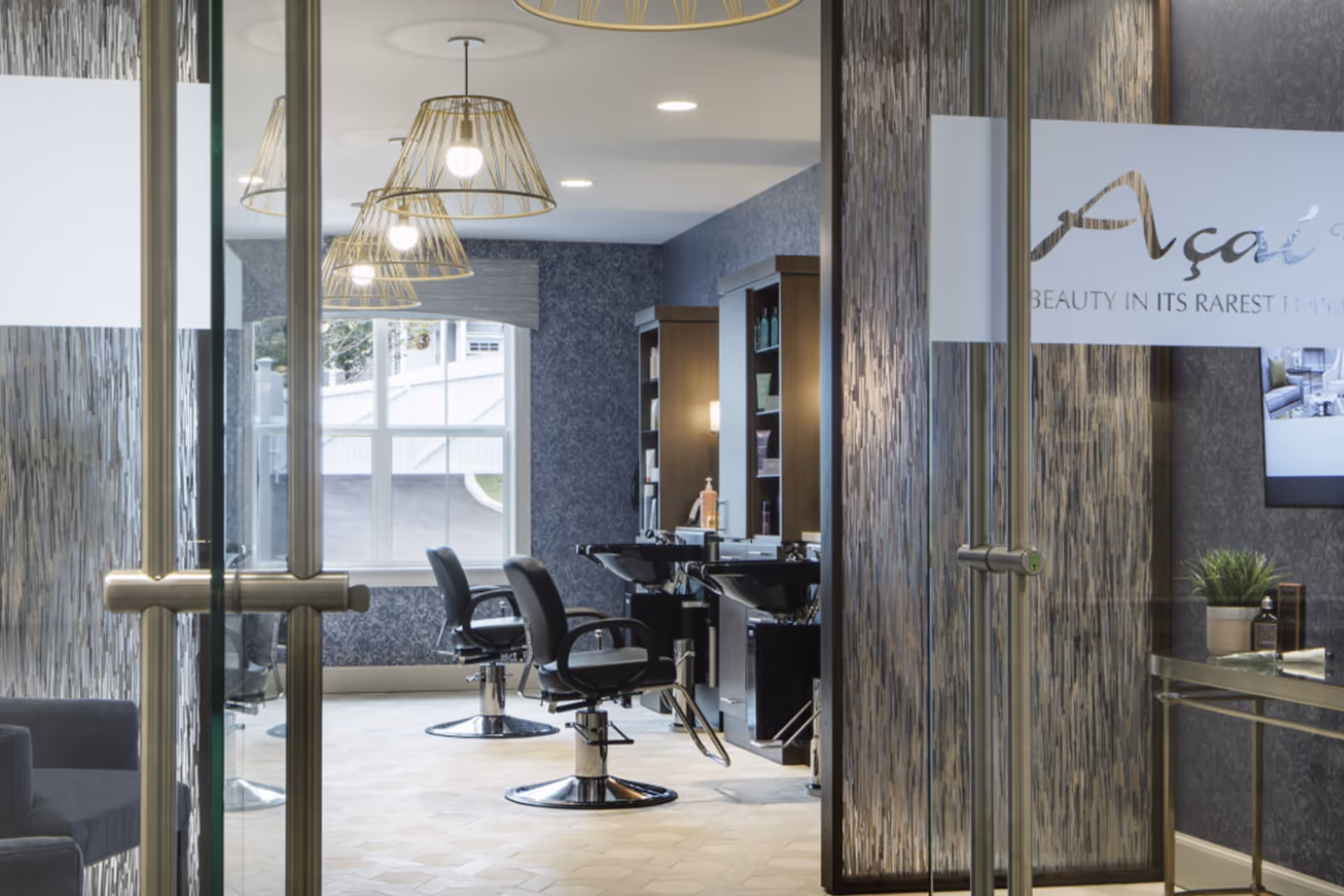 Interior view of a modern hair salon area with black styling chairs, wash basins, shelves with hair products, and pendant lights hanging from the ceiling. The room has a large window letting in natural light and glass doors at the entrance.