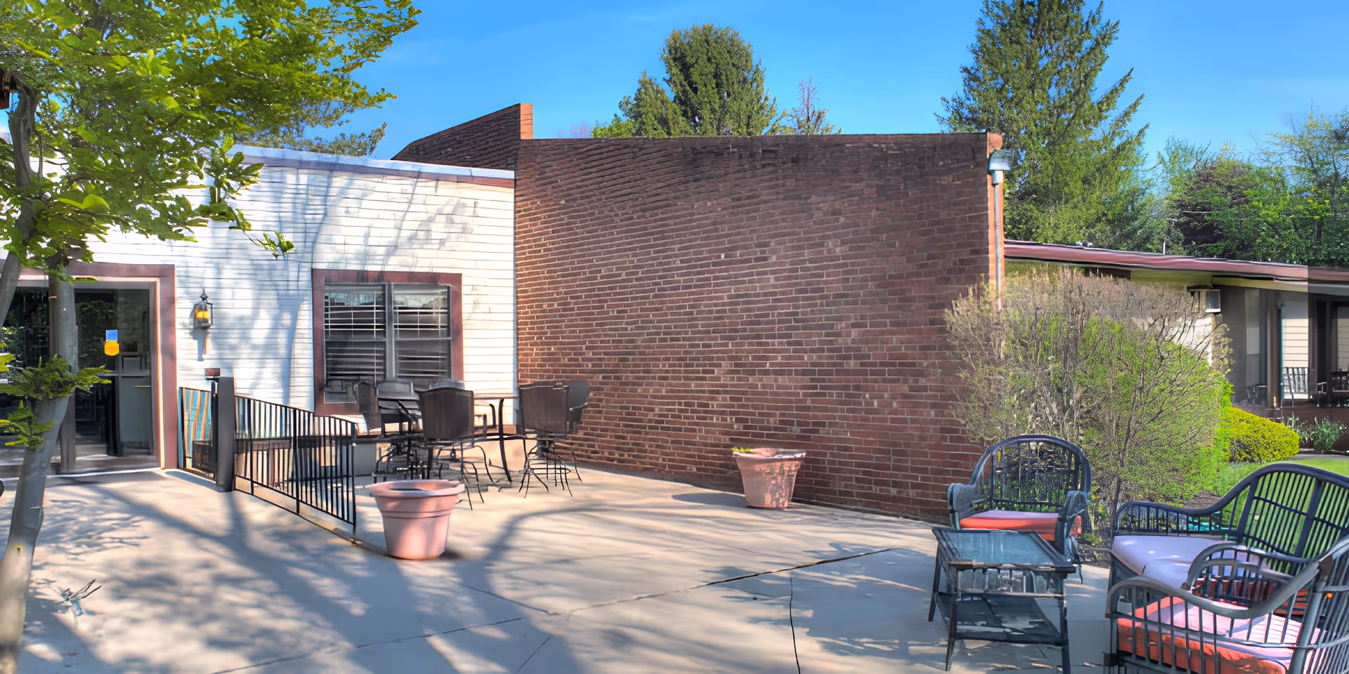 Outdoor patio area at Springfield Assisted Living with metal chairs and tables, potted plants, and surrounding greenery including trees and bushes under a clear blue sky.