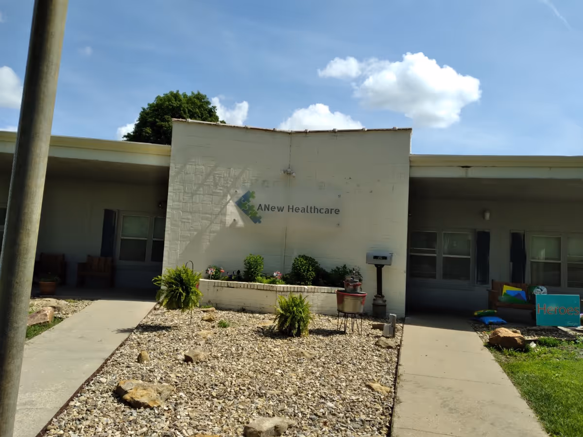 Exterior view of ANEW Healthcare building with a white brick facade, a small garden area with plants and rocks in front, and a clear blue sky with some clouds above.