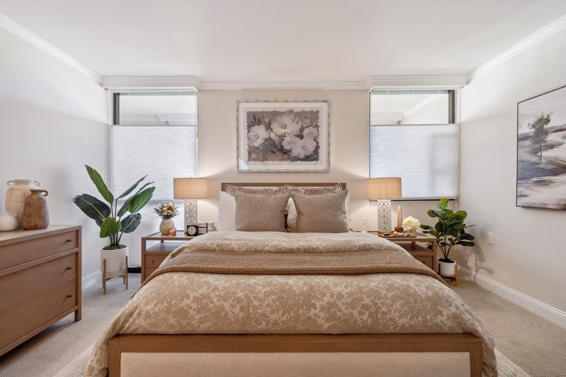 A neatly made bed with beige floral bedding and two large pillows in a bright bedroom. On either side of the bed are wooden nightstands with table lamps, decorative items, and plants. There is a wooden dresser with vases on the left side and framed artwork on the walls. Two windows with white blinds flank the bed.