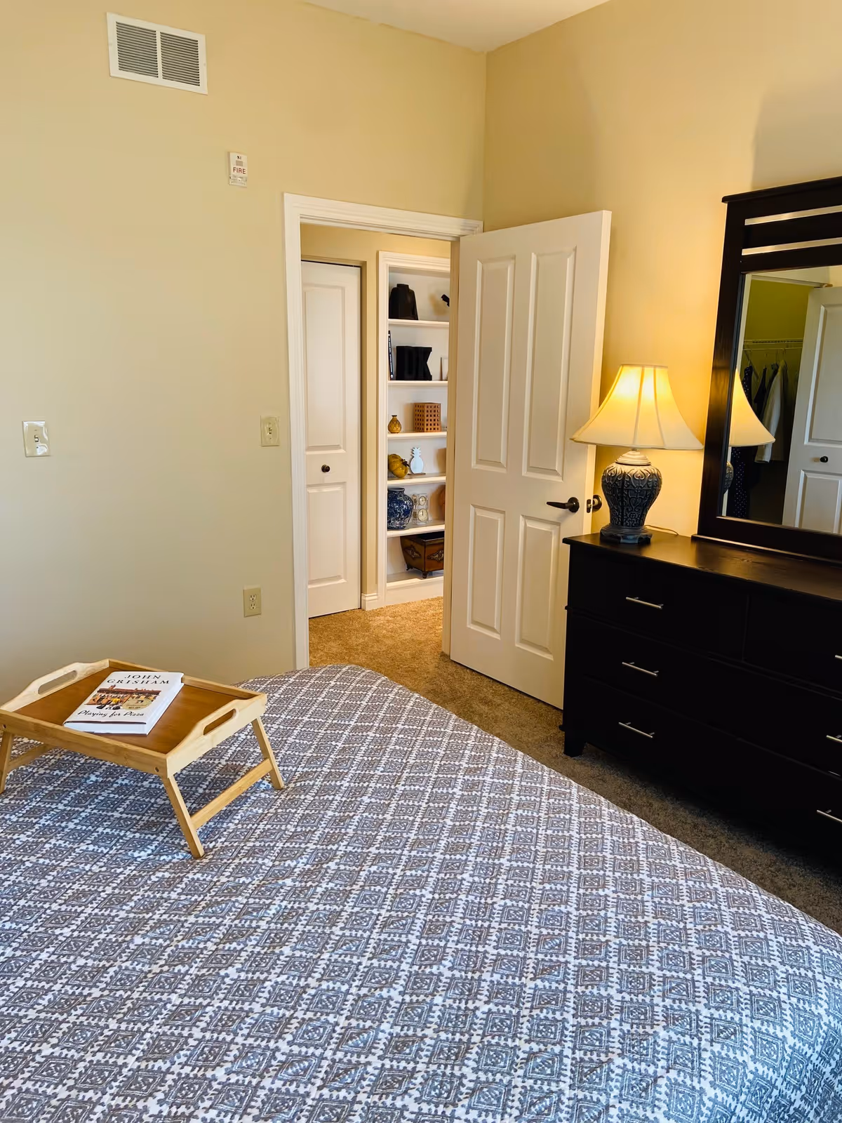 A bedroom with a patterned bedspread and a wooden tray holding a book. There is a dark wooden dresser with a lamp and mirror on top. The door is open, showing a hallway with shelves holding decorative items and a closet with hanging clothes.