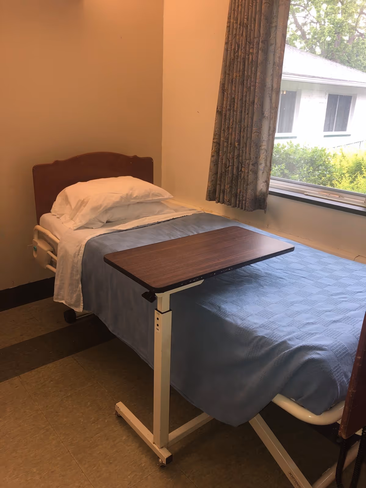 A single bed with a wooden headboard, white pillow and sheets, and a blue blanket in a small room. There is an adjustable overbed table positioned over the bed. A window with floral curtains shows greenery and part of another building outside.
