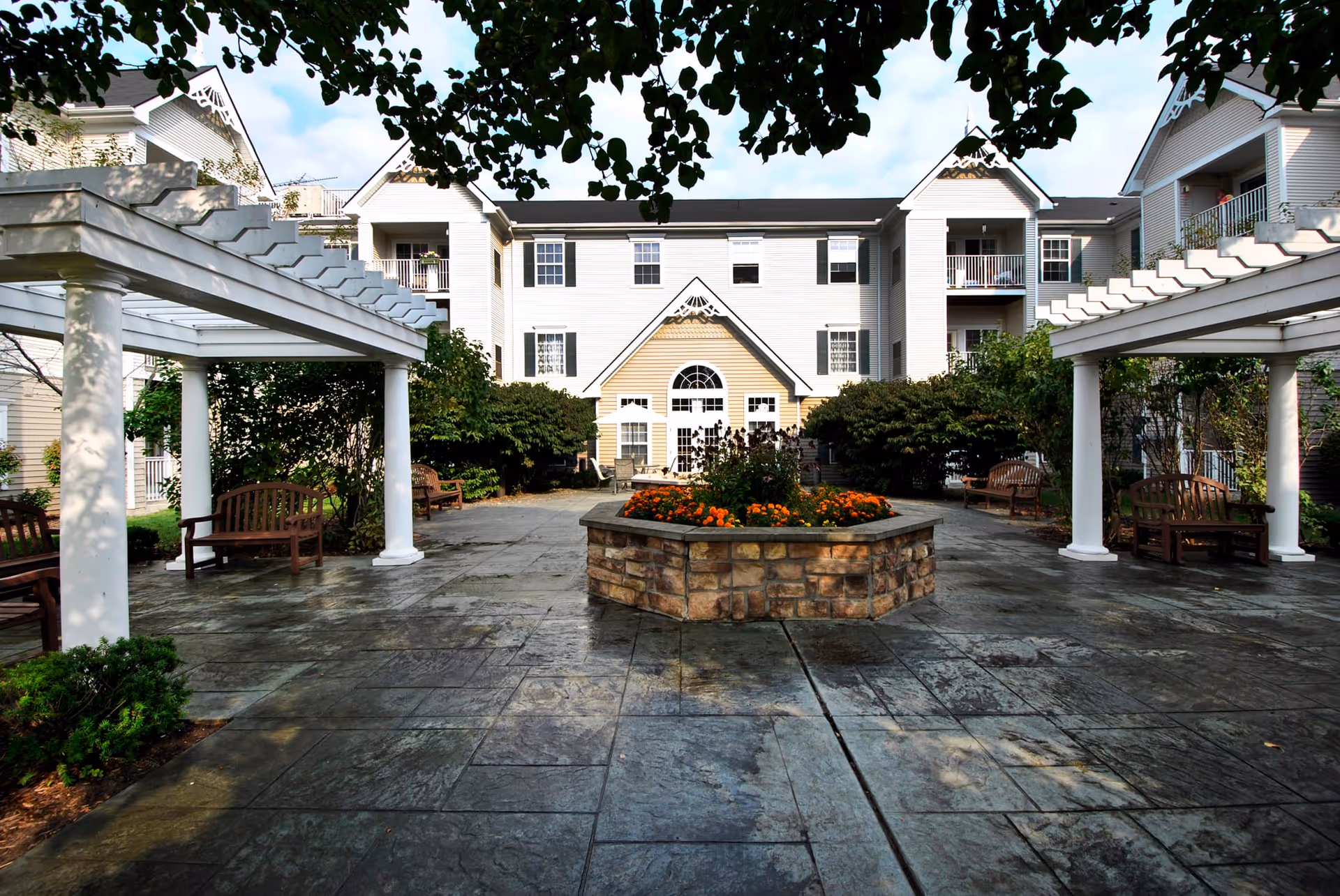 Outdoor courtyard area of a senior living facility with a stone-paved ground, a central raised flower bed with orange flowers, wooden benches under white pergolas, and a multi-story building with balconies and windows in the background.