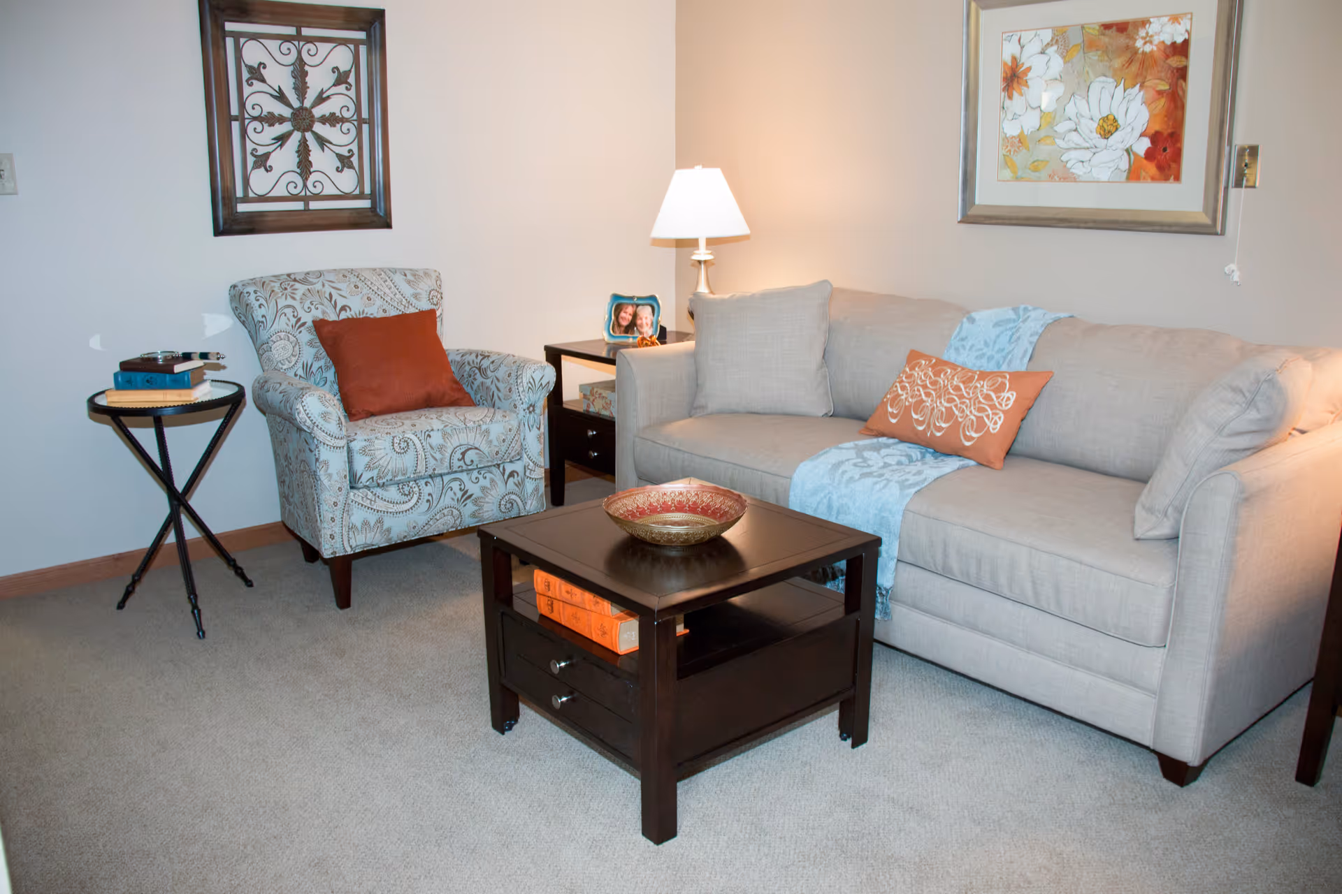 A cozy living room with a beige sofa adorned with decorative pillows and a blue throw blanket. Next to the sofa is a patterned armchair with a rust-colored pillow. A dark wooden coffee table with a decorative bowl and books underneath sits in front of the sofa. The walls are decorated with framed artwork and a table lamp on a side table provides warm lighting.