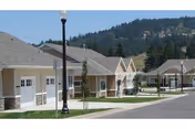A row of single-story residential buildings with beige siding and stone accents, situated along a paved road with a streetlamp and young trees. Hills covered with trees are visible in the background under a clear sky.