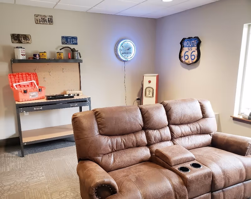 A cozy room with a brown leather reclining sofa in the foreground. On the back wall, there is a workbench with a red toolbox and various tools and containers. Above the workbench are vintage license plates. A neon clock and a Route 66 sign are mounted on the adjacent walls. A window on the right lets in natural light.