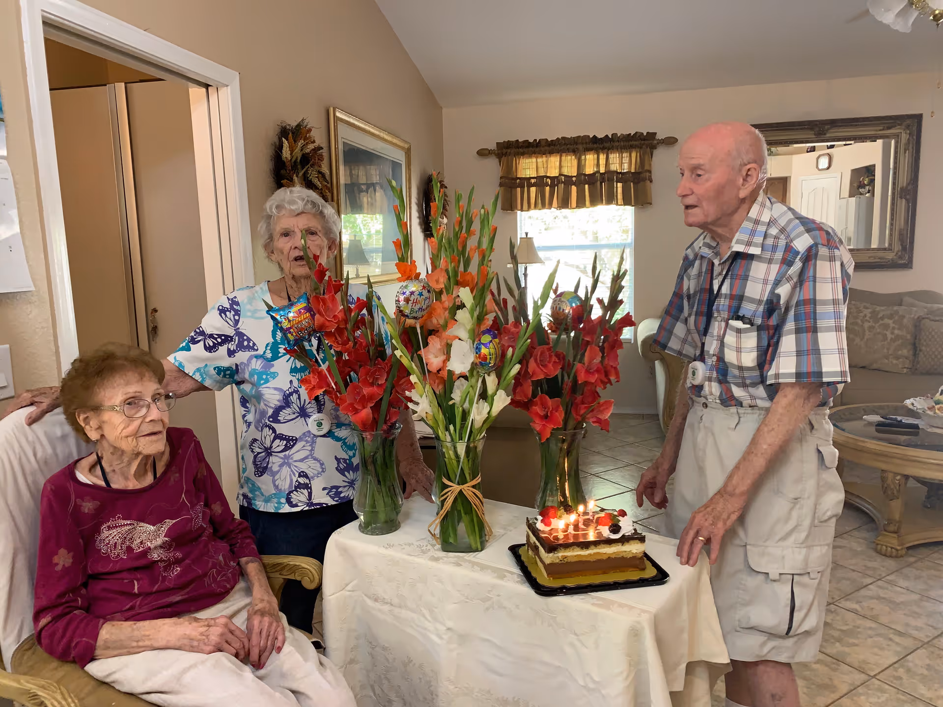 Three elderly people in a living room setting celebrating a birthday with a cake on a table decorated with three vases of red and orange flowers. One woman is seated, another woman stands behind her, and a man stands on the opposite side of the table. The room has a window with curtains, a mirror on the wall, and a couch in the background.