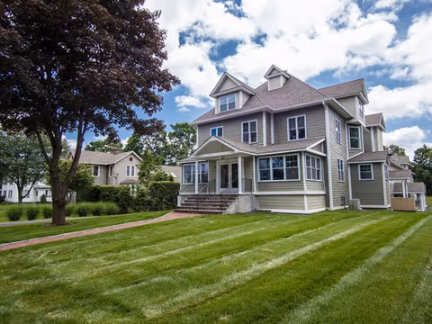 A large, multi-story residential building with beige siding and white trim, surrounded by a well-maintained green lawn and trees under a partly cloudy sky.