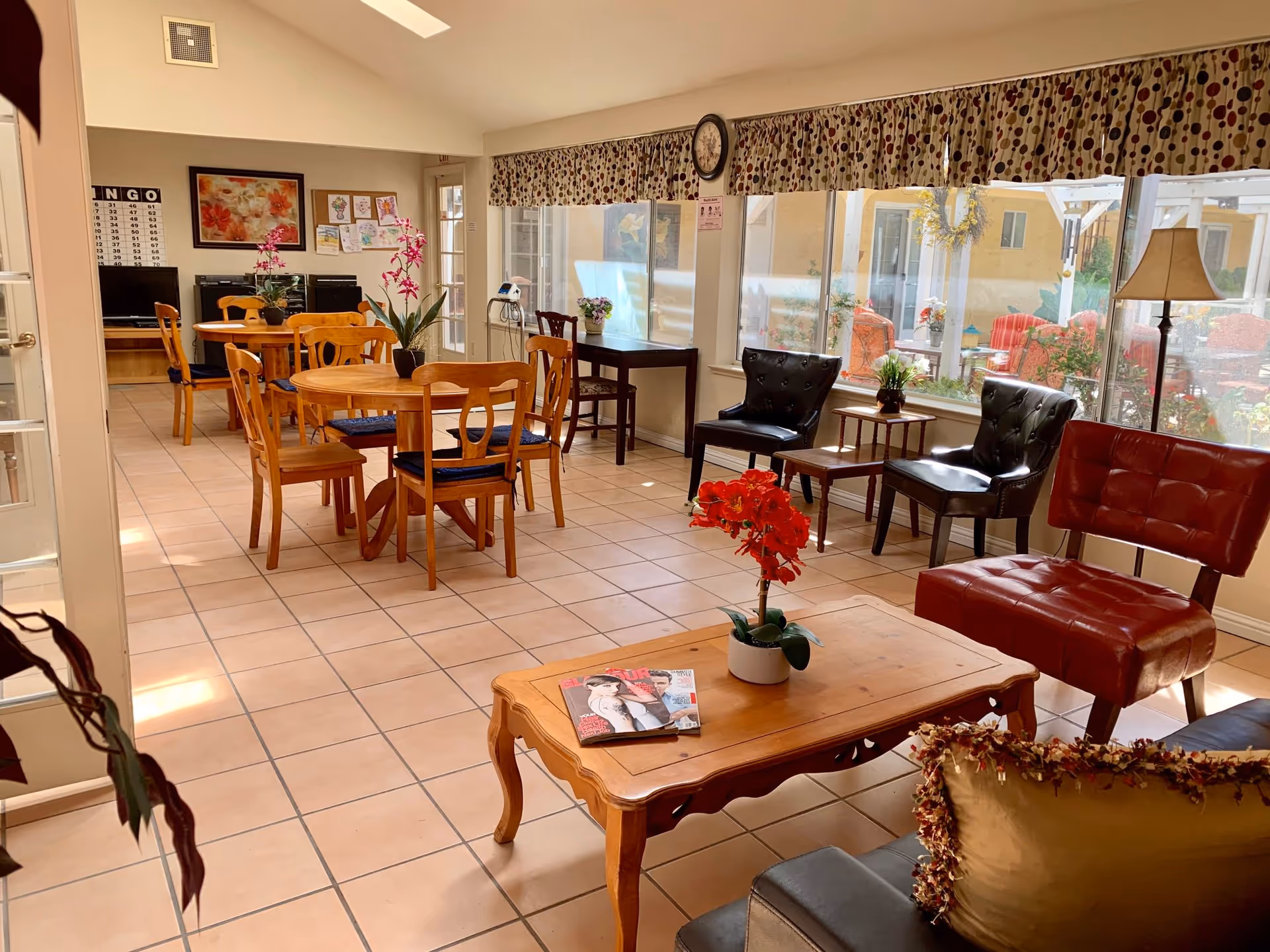 Sunlit communal dining and lounge area with wooden tables and chairs, armchairs, a coffee table and large windows overlooking a courtyard.