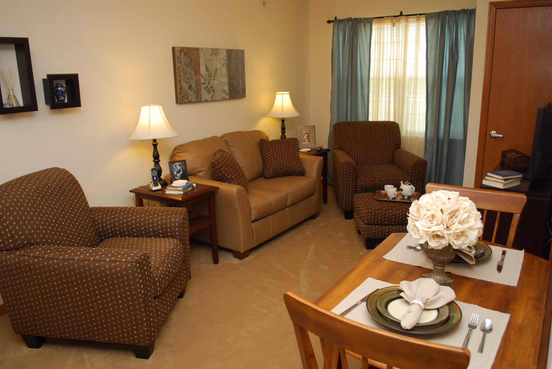A cozy living room with a tan leather loveseat, two brown patterned armchairs, and a wooden dining table set for two with plates, napkins, and a vase of white flowers. The room has beige carpet, two table lamps, wall art, a window with blue curtains, and a TV on a stand with books nearby.