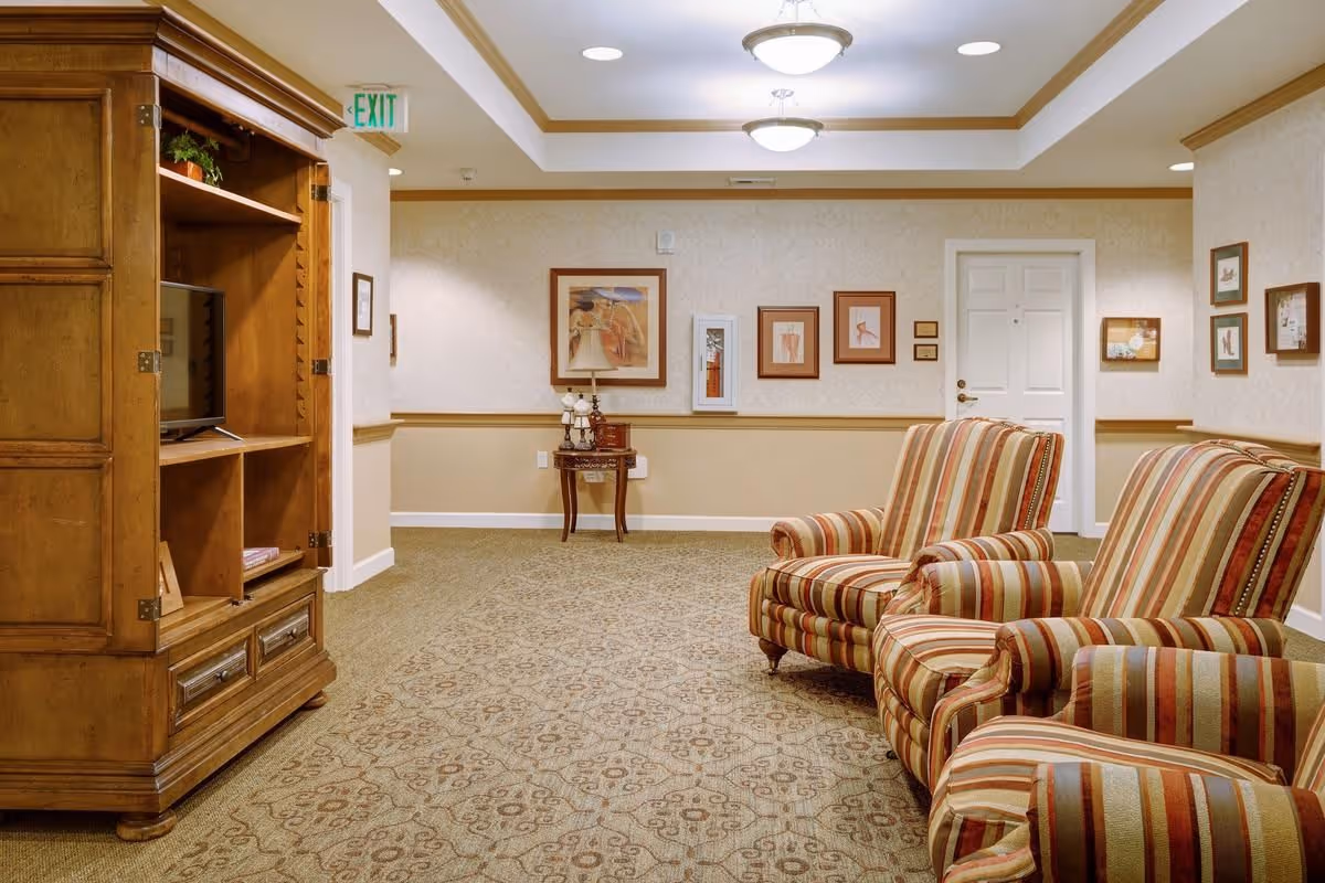 A senior living common room with striped armchairs, a wooden entertainment cabinet, framed artwork, and patterned carpet.