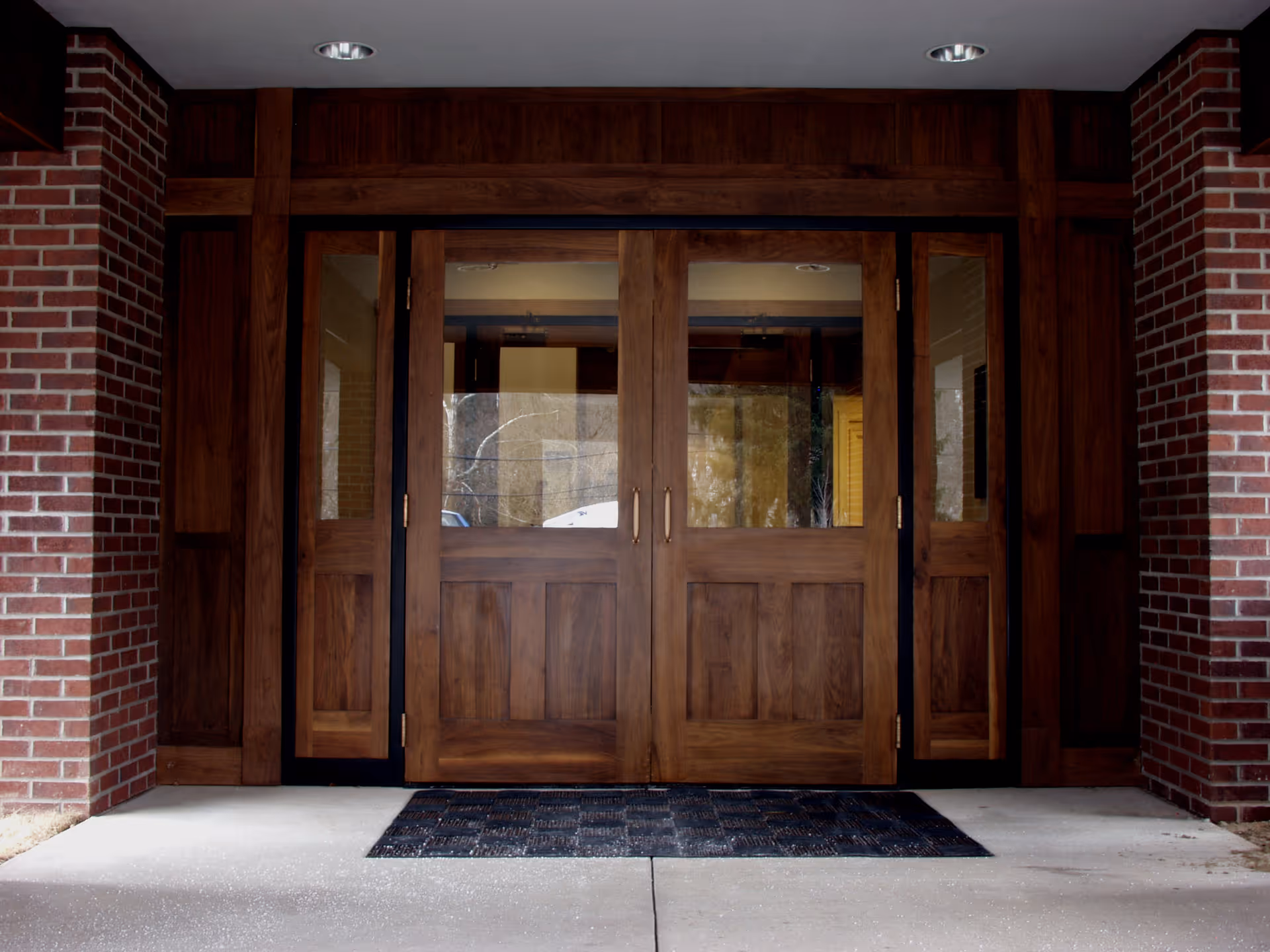 Entrance to a building with double wooden doors featuring glass panels, flanked by brick walls and a dark doormat on the concrete floor.
