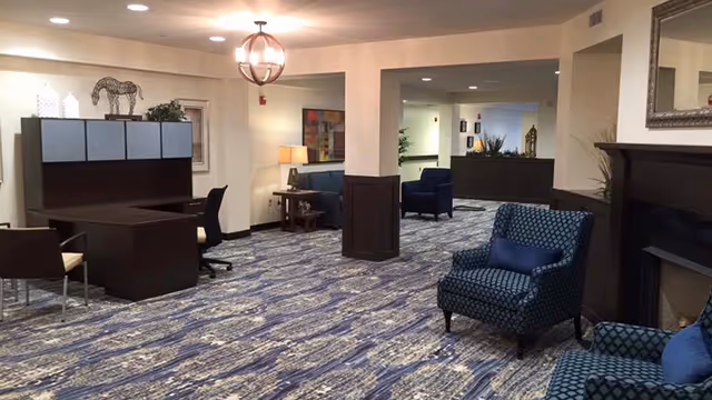 A spacious interior room with patterned blue and beige carpet, featuring several seating areas with blue upholstered armchairs and a dark wooden desk with chairs. The room has warm lighting from ceiling fixtures and table lamps, with decorative elements including a zebra sculpture on a cabinet and framed artwork on the walls.